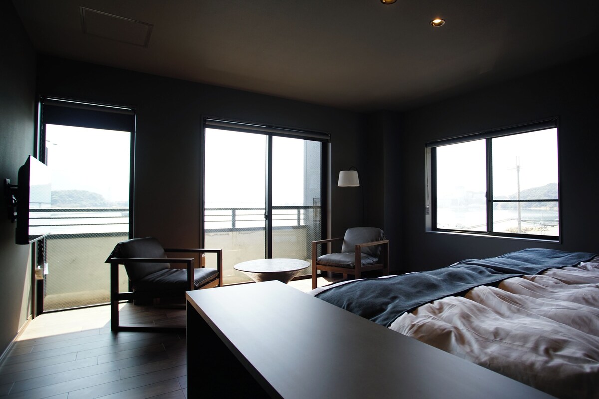 Une chambre à coucher avec un lit, une chaise, une petite table et de grandes fenêtres.