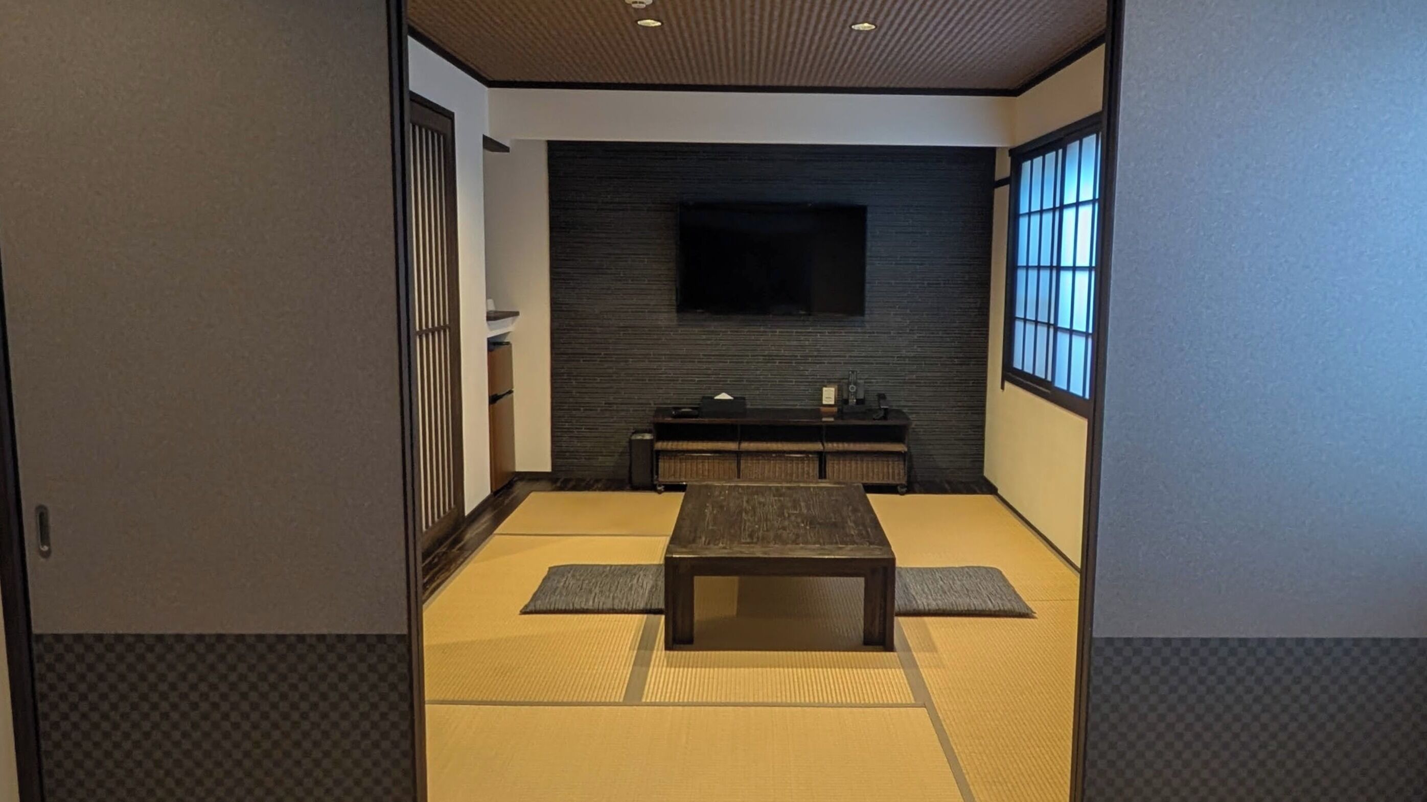 Ein traditioneller japanischer Raum mit Tatami-Bodenbelag, einem niedrigen Tisch, einem an der Wand montierten Fernseher und Schiebetüren.