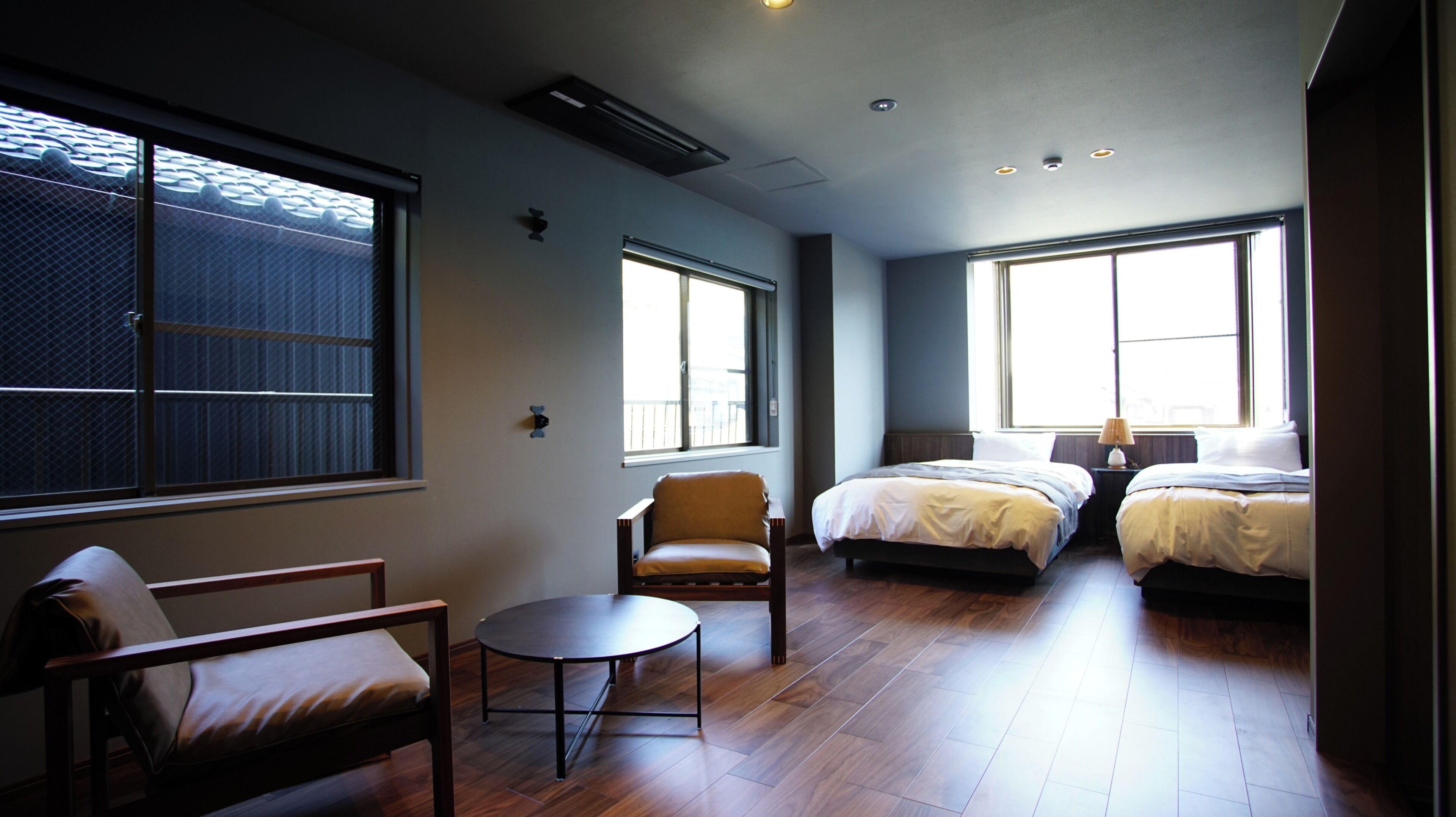 Ein Hotelzimmer mit zwei Betten, einem Sessel, einem kleinen Tisch und großen Fenstern.