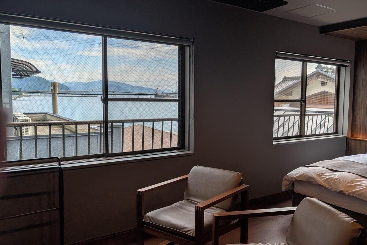 A hotel room with a bed, two chairs, and large windows offering a view of a body of water and buildings.