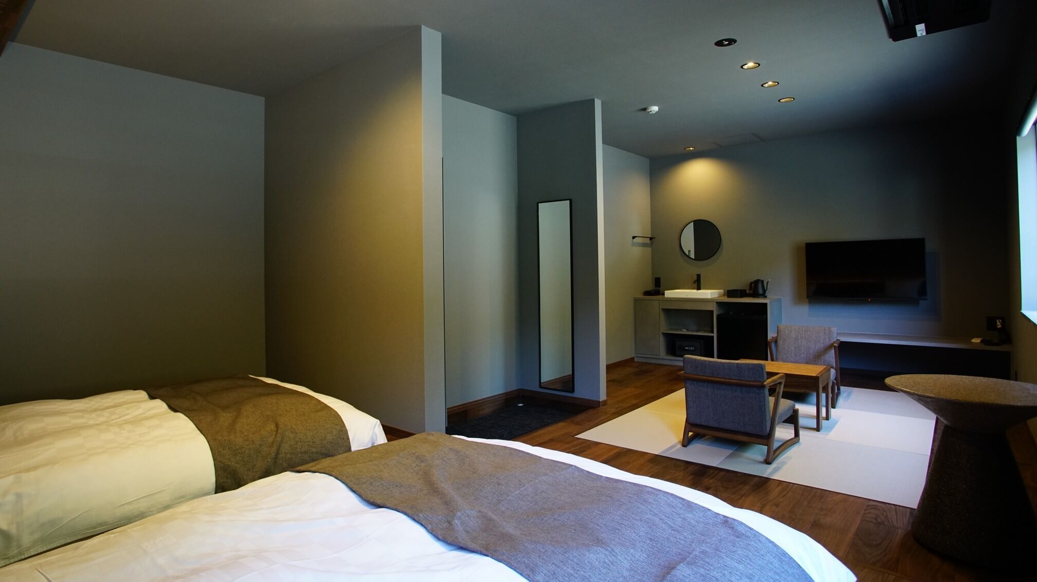 Ein modernes Hotelzimmer mit Bett, Schreibtisch, Sessel, kleinem Tisch, Spiegel, Fernseher und Waschbecken.
