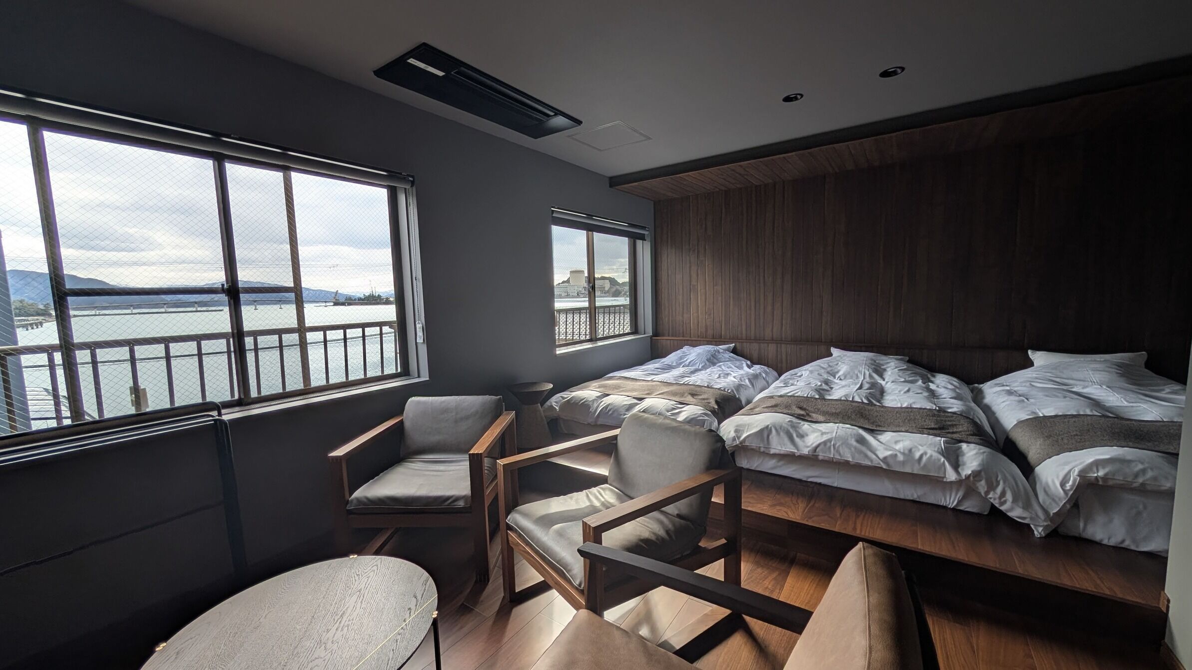 Ein modernes Schlafzimmer mit einem großen Bett, zwei Sesseln, einem kleinen Tisch und großen Fenstern mit Blick auf ein Gewässer und ferne Hügel.