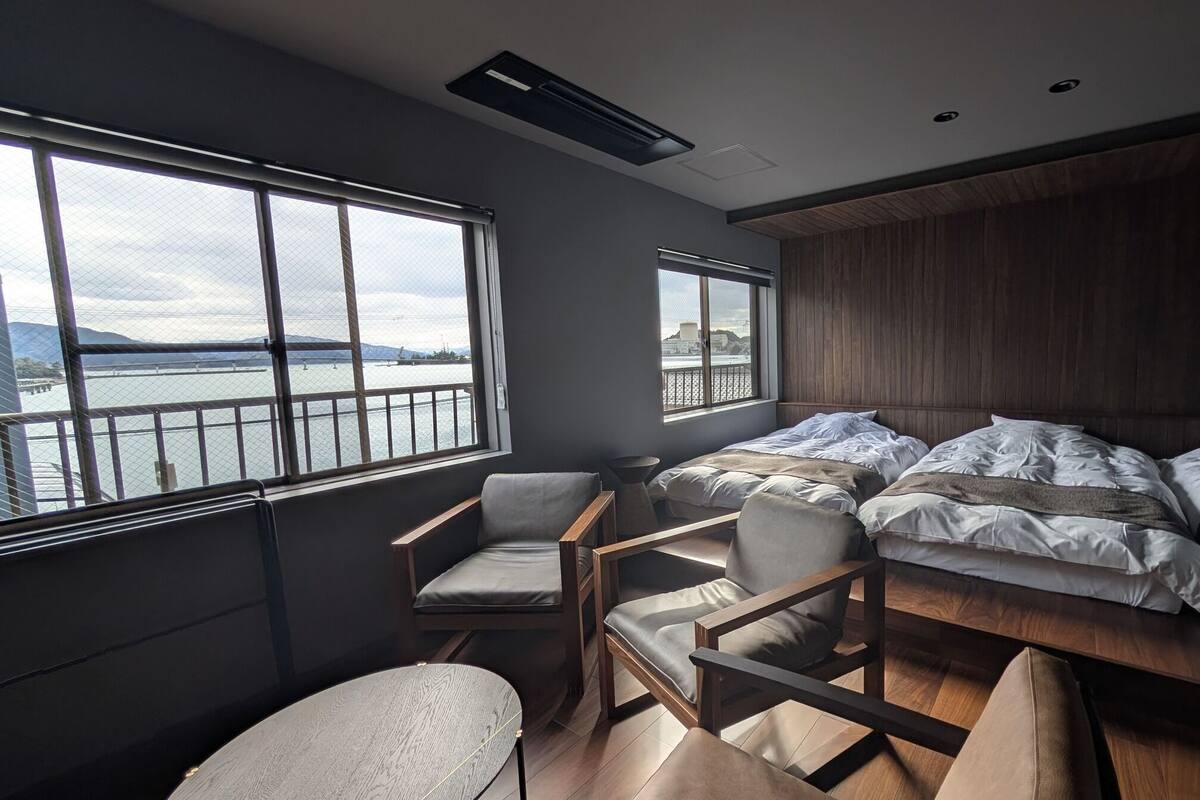 Camera moderna con un letto grande, due poltrone, un tavolino e ampie finestre con vista su un corpo d'acqua e colline lontane.