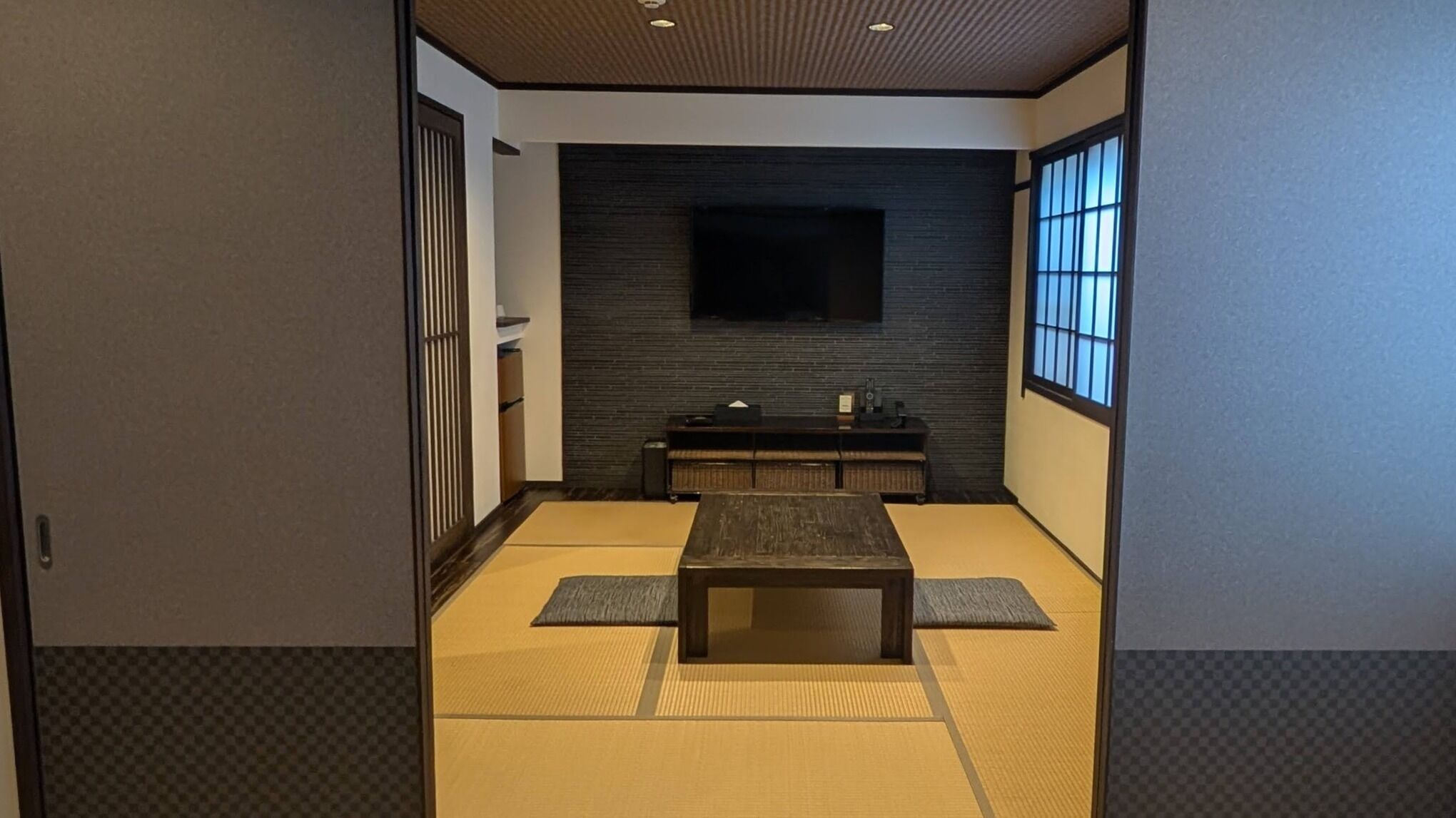Ein traditioneller japanischer Raum mit Tatami-Boden, einem niedrigen Tisch, einem an der Wand montierten Fernseher und Schiebetüren.