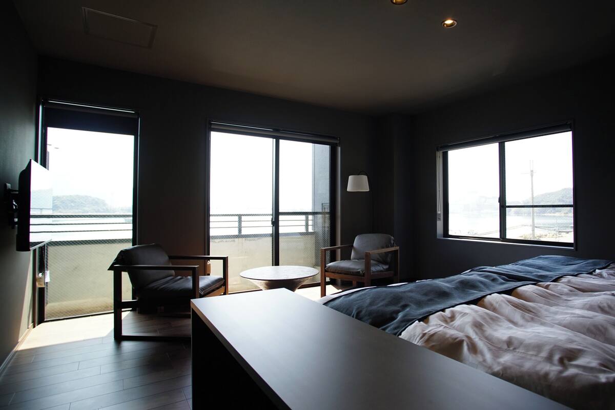 Una camera da letto con un letto, una sedia, un tavolino e ampie finestre.