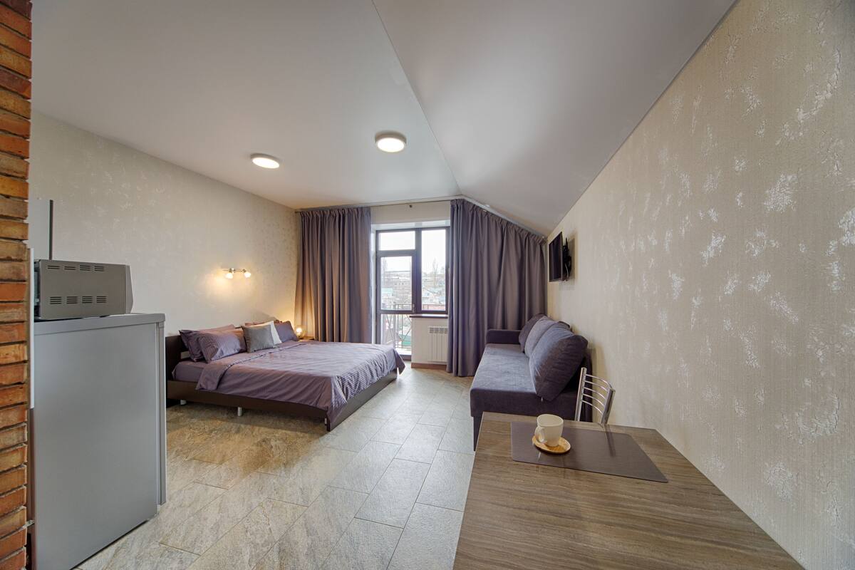 A compact hotel room with a bed, a sofa, a desk with a cup, and a TV on the wall.