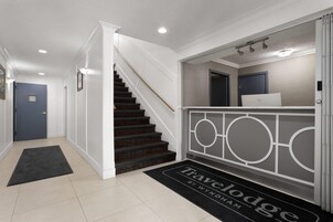 Un pasillo limpio y moderno con una escalera que conduce a una plataforma. Se ve un ordenador portátil sobre una encimera, y un felpudo con la inscripción "Travelodge by Wyndham" en la entrada.