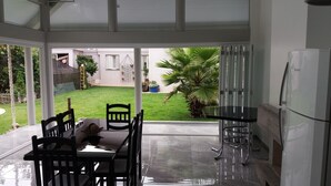 A covered patio area with a dining table and chairs, a refrigerator, and a view of a garden with a palm tree and a house in the background. - Casa da Piscina Bento Gonçalves (Bento Gonçalves)