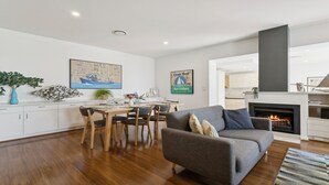 A modern living room with a fireplace, a dining area with wooden furniture, and a kitchen in the background. - Koonya Sur Mer Coastal Sorrento Escape (Sorrento)