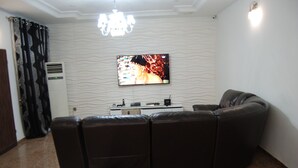 A living room with a sectional sofa, a flat-screen TV mounted on the wall, a ceiling light fixture, and a wall-mounted air conditioner. - Kings Court Apartment (Abuja)