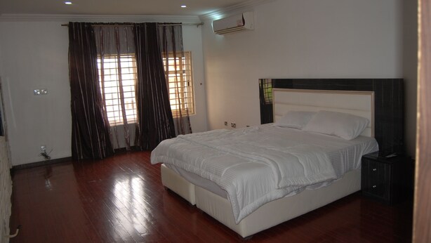 A bedroom with a large bed, a bedside table, a wardrobe, and an air conditioning unit. - Kings Court Apartment (Abuja)