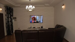 A living room with a flat-screen TV mounted on the wall, a brown leather sofa, a chandelier, and a patterned curtain. - Kings Court Apartment (Abuja)