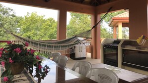 A covered outdoor dining area with a table, chairs, a hammock, and a grill.