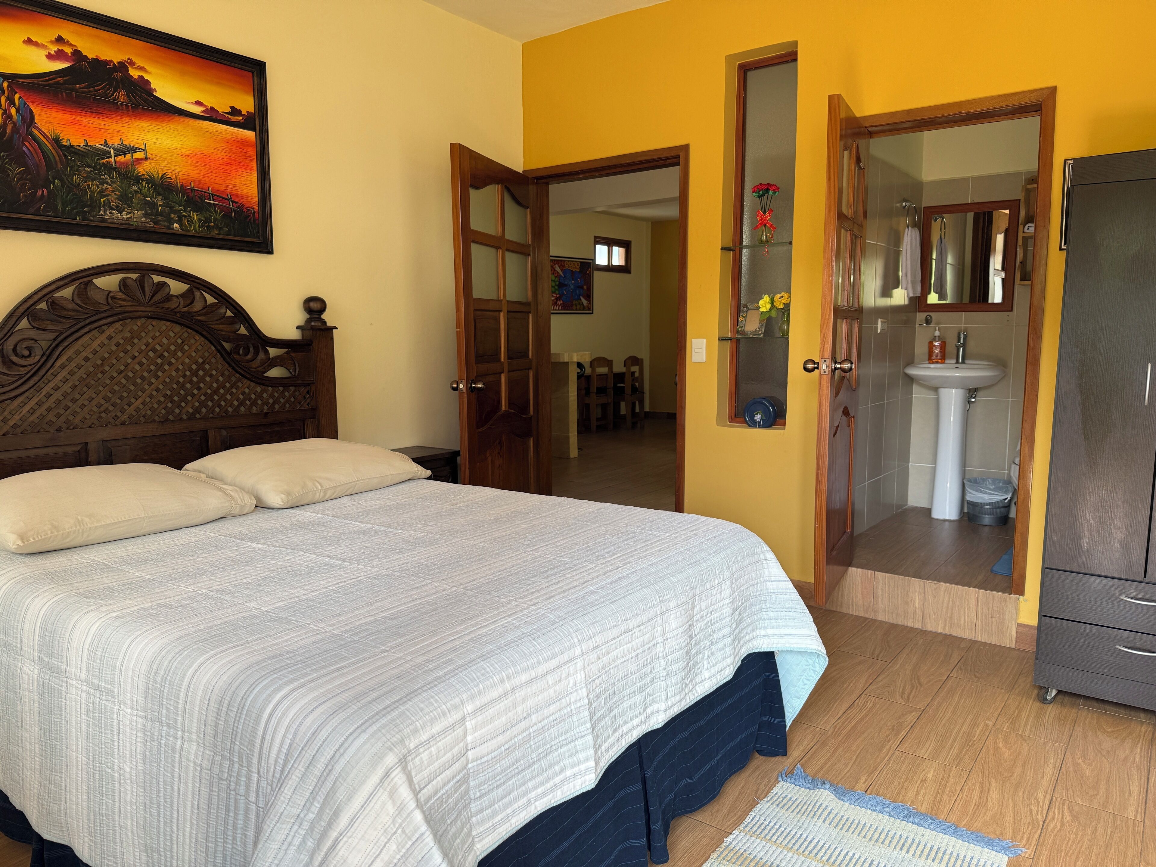 Un dormitorio con cama, cabecera de madera, un cuadro, baño con lavamanos y espejo, y un armario.