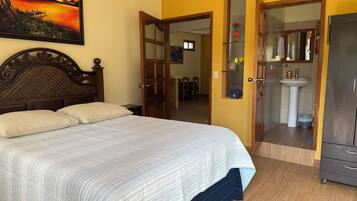 Un dormitorio con cama, cabecera de madera, un cuadro, baño con lavamanos y espejo, y un armario.