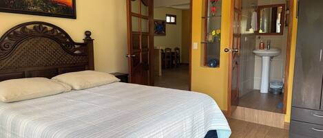 A bedroom with a bed, a wooden headboard, a painting, a bathroom with a sink and mirror, and a wardrobe.