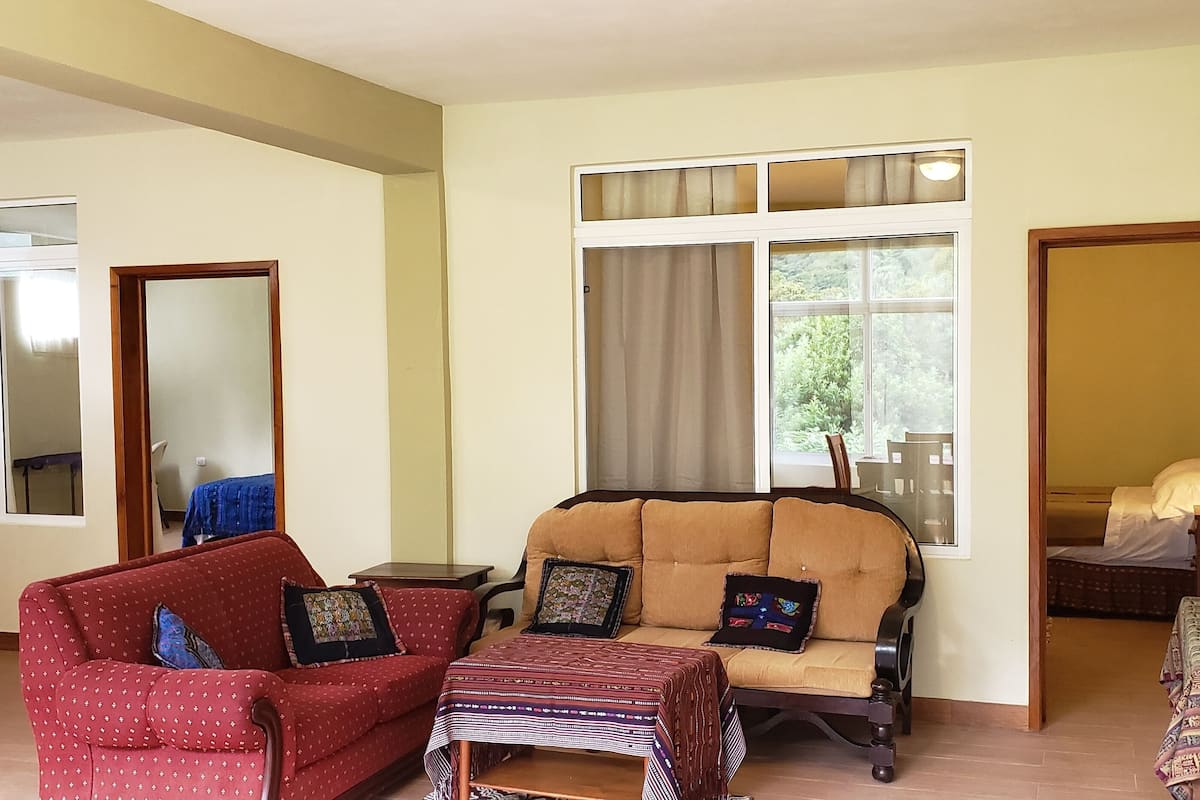 Una sala de estar con dos sofás, una mesa de centro y un televisor. Se ve una ventana con cortinas, una puerta que da a otra habitación y una mesita con una lámpara.