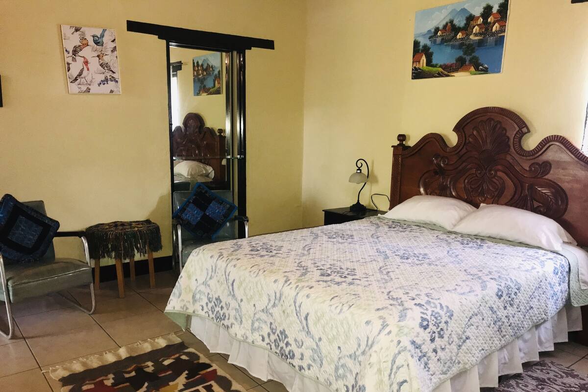 Un dormitorio con una cama grande, una silla, una mesita y una puerta que da a otra habitación.