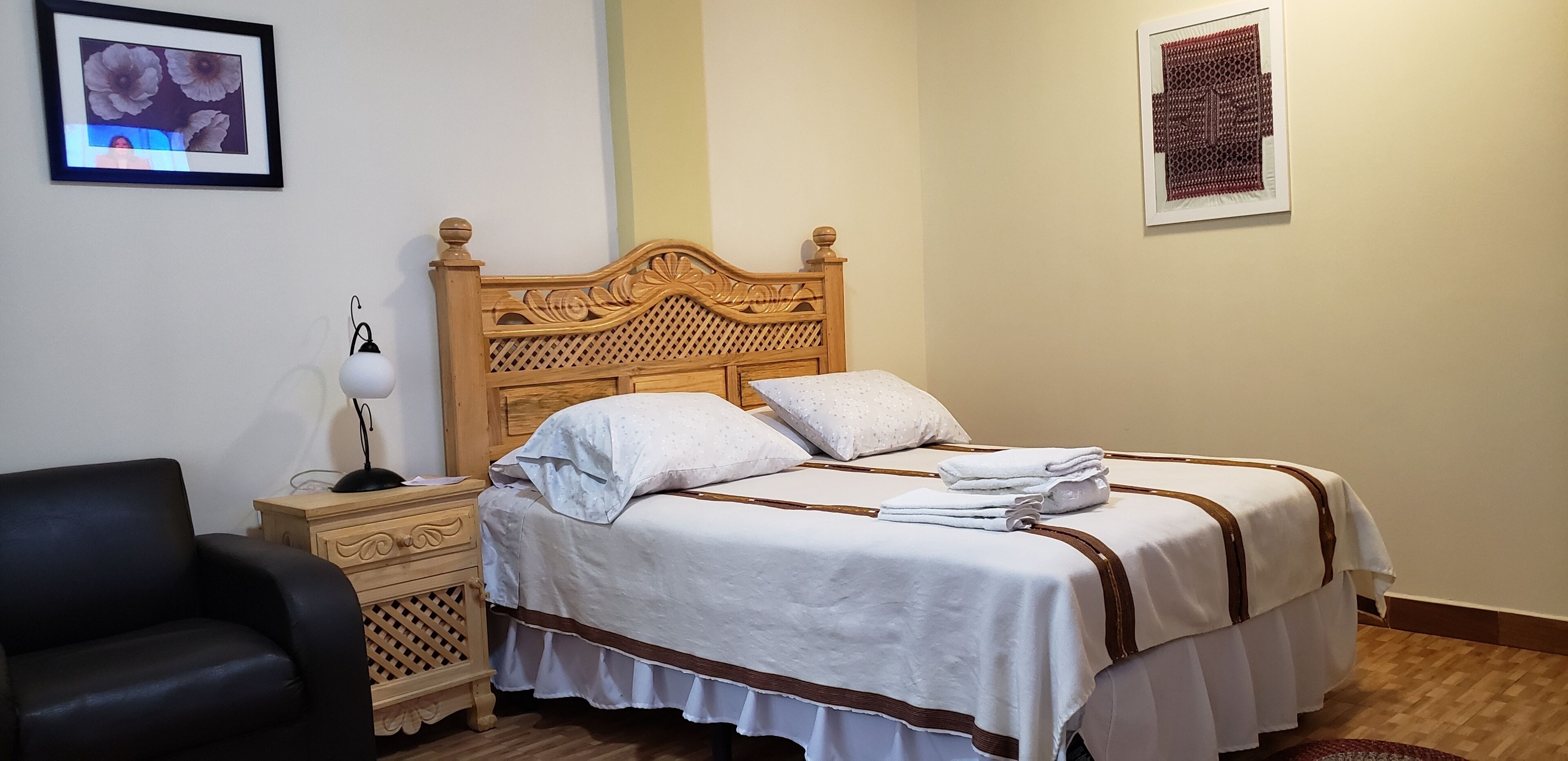A neatly made bed with a wooden headboard, a black chair, a bedside table with a lamp, and two framed pictures on the walls.