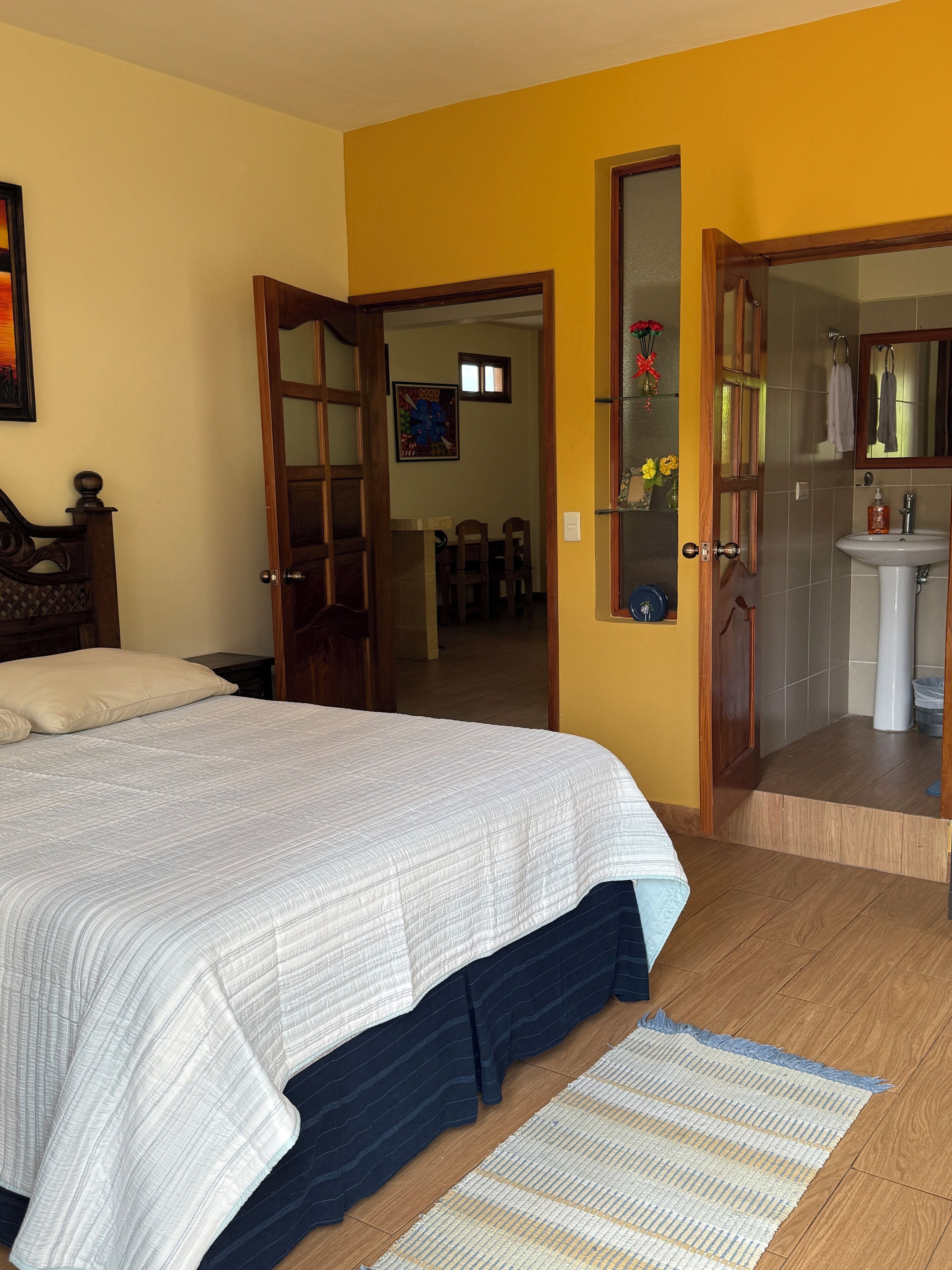 Un dormitorio con cama, una puerta de madera, un baño con lavamanos y espejo, y un comedor con sillas y mesa.