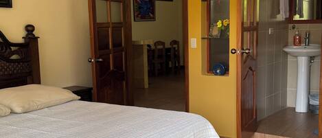 Un dormitorio con cama, una puerta de madera, un baño con lavamanos y espejo, y un comedor con sillas y mesa.