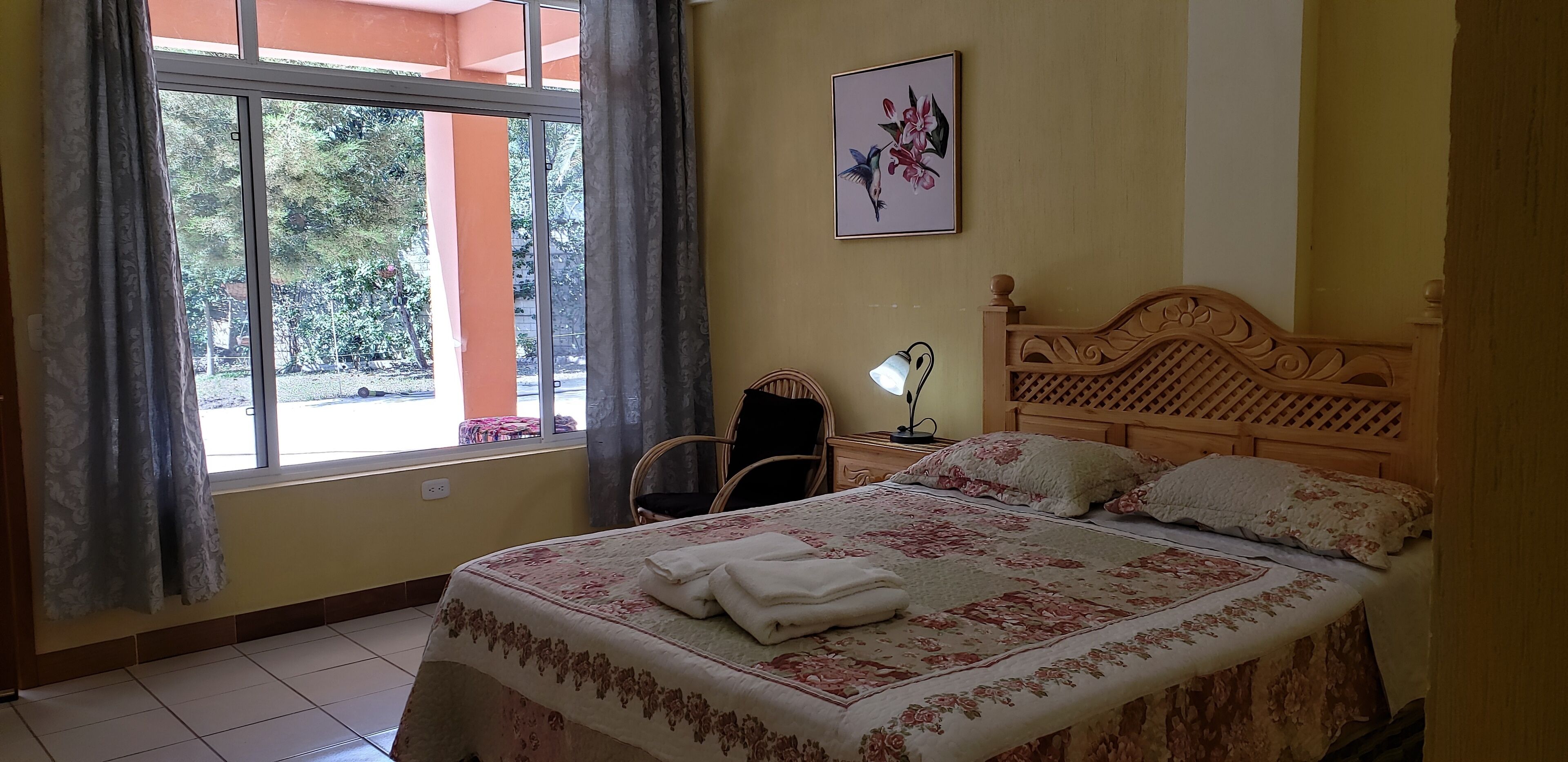 A bedroom with a bed, a nightstand, a lamp, a chair, and a window with curtains.