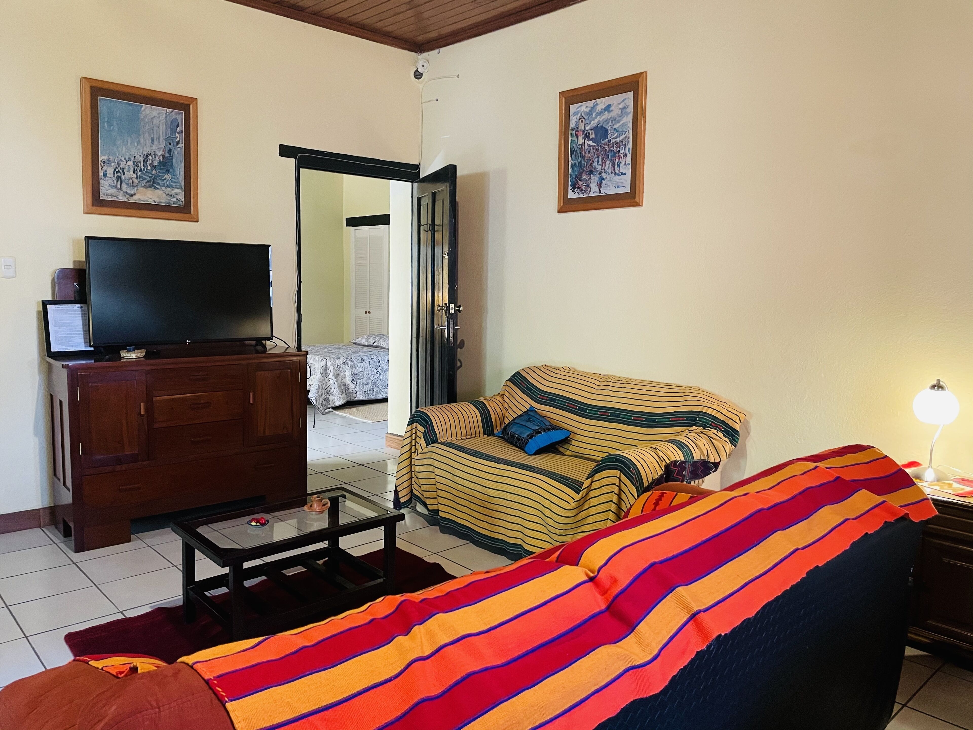 A living room with a sofa, a television on a wooden cabinet, and a bed in the background.
