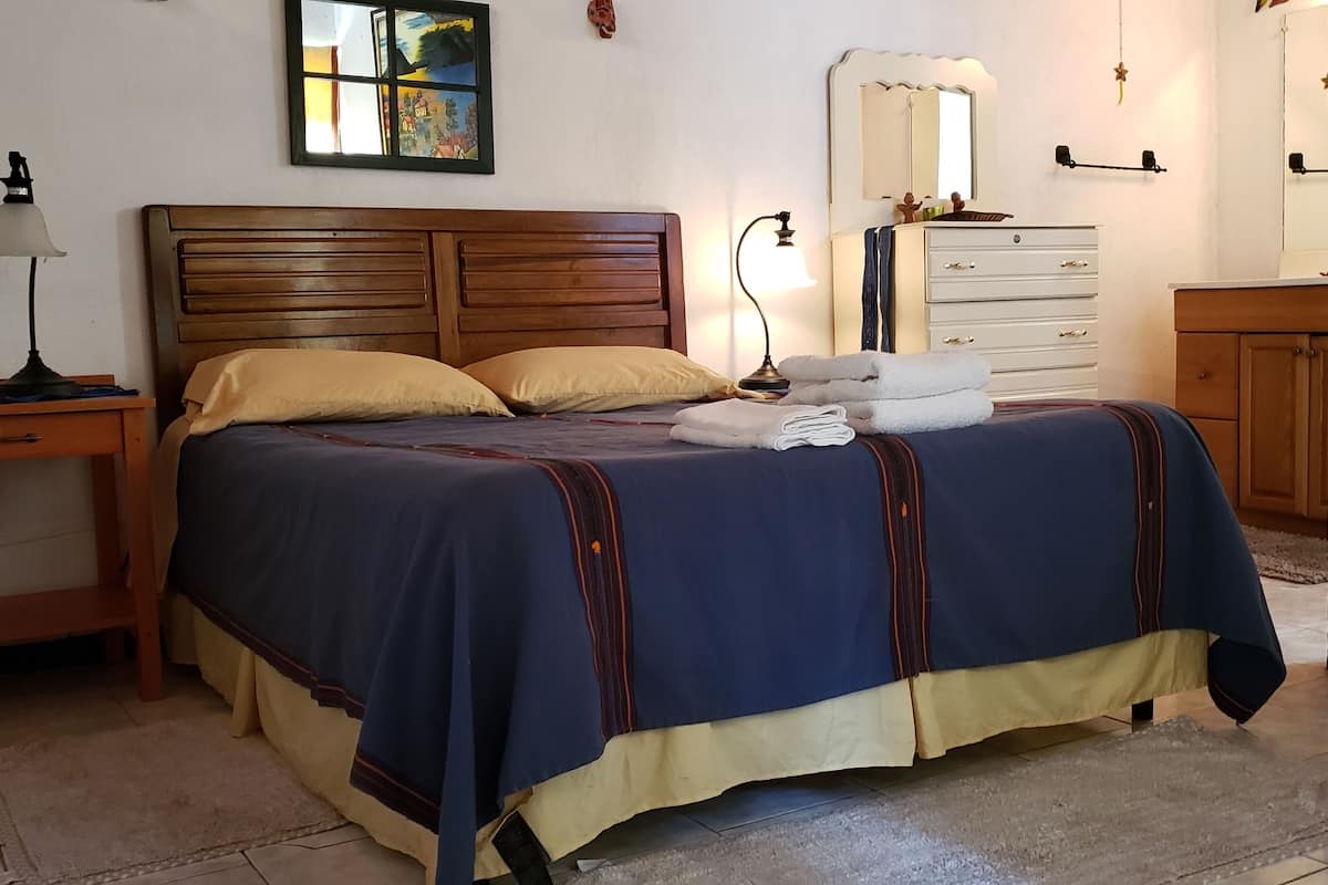 Una cama bien hecha con colcha azul y amarilla, cabecera de madera, mesita de noche con lámpara, cómoda con espejo y un cuadro en la pared.