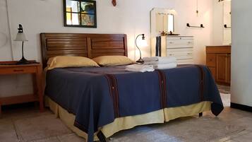 Una cama bien hecha con colcha azul y amarilla, cabecera de madera, mesita de noche con lámpara, cómoda con espejo y un cuadro en la pared.