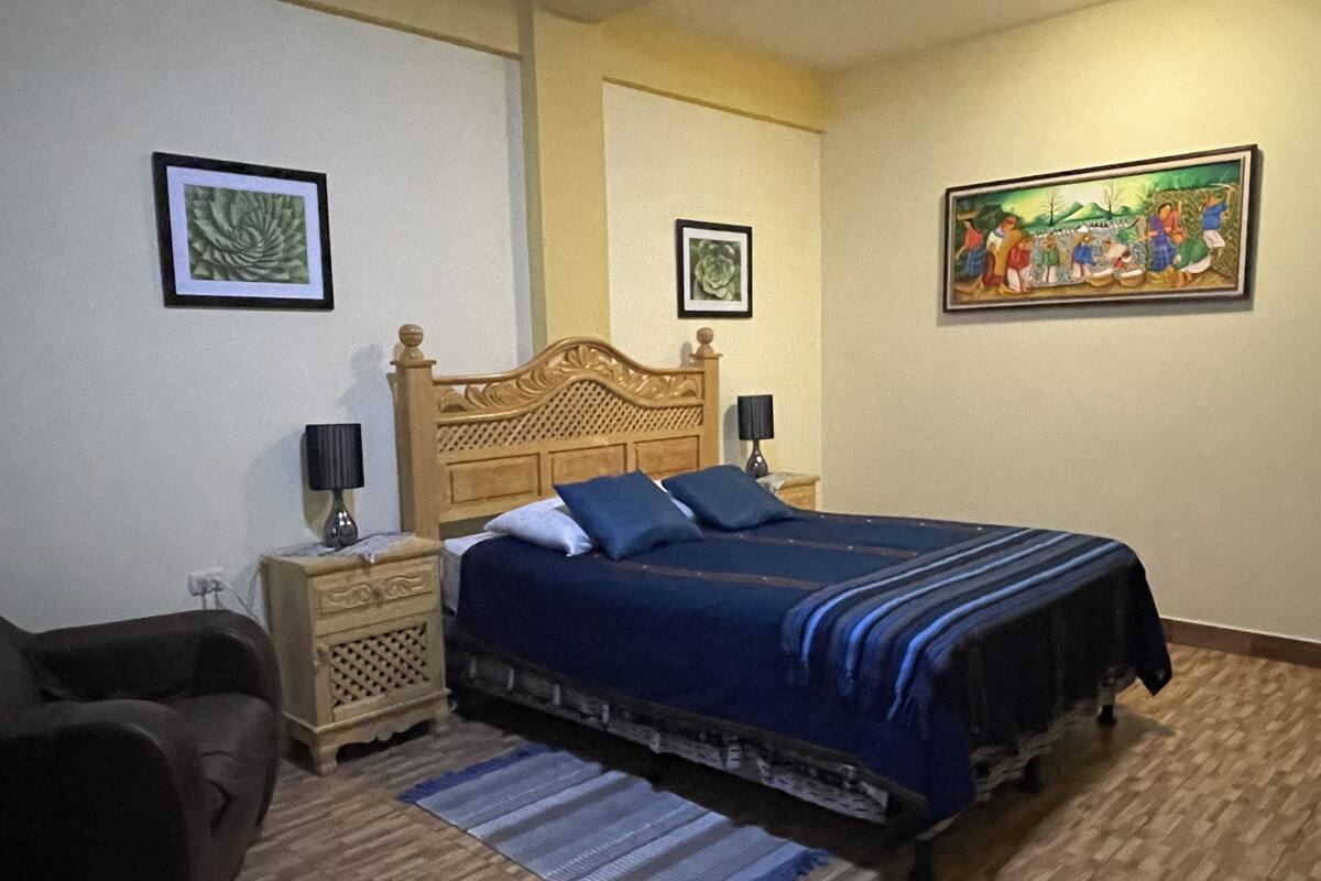 Un dormitorio con una cama de madera, dos mesitas de noche, una mesita auxiliar, un tapete azul y negro a rayas y dos cuadros enmarcados en las paredes.