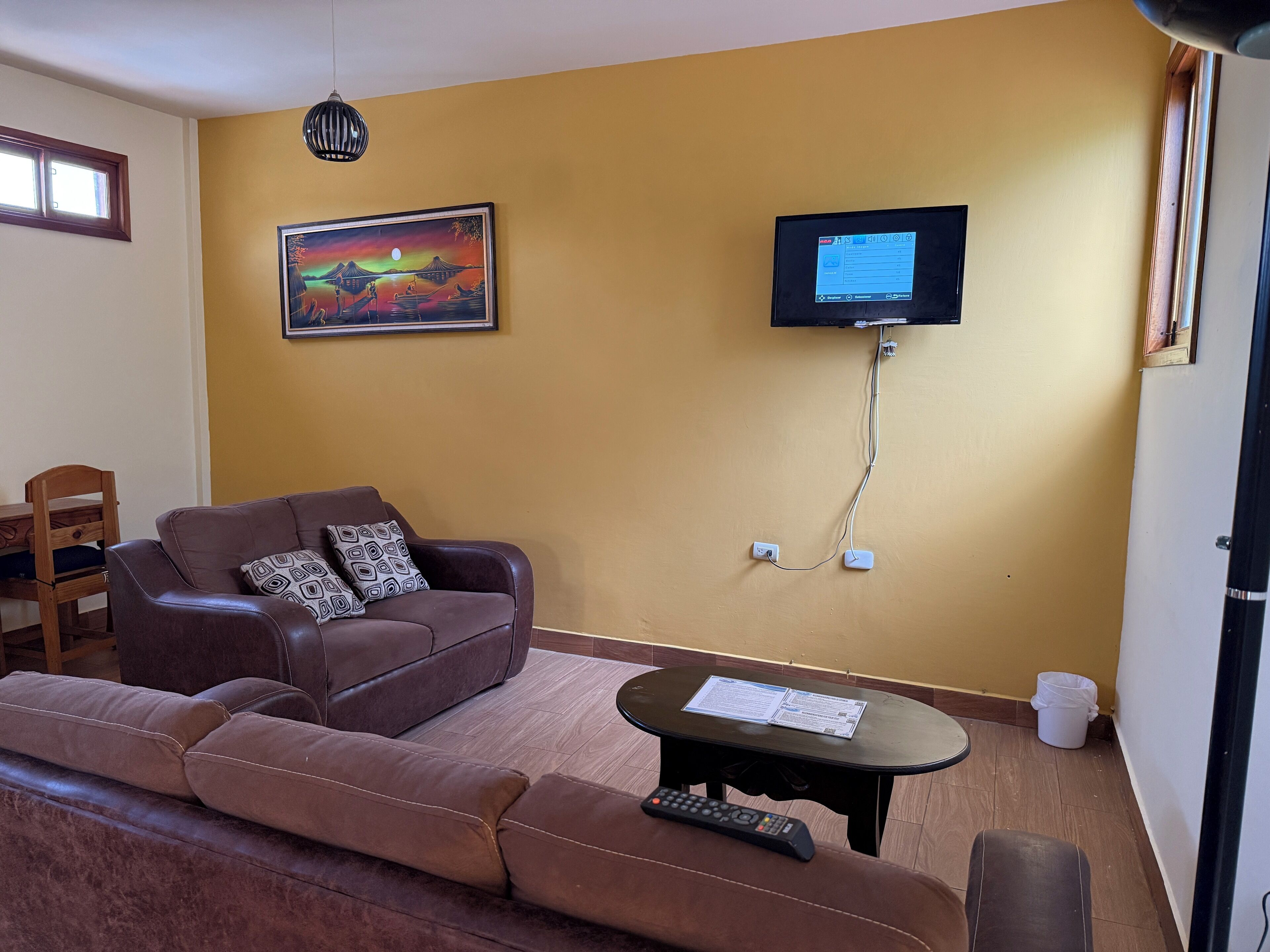 Ein Wohnzimmer mit einem braunen Ledersofa, einem kleinen Tisch mit Papieren, einem Wand-Fernseher und einem Bild an der Wand.
