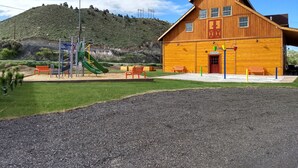 Un edificio de madera con techo inclinado, un parque infantil con toboganes y columpios, y un césped delantero.