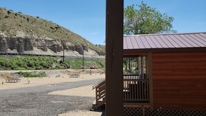 Una cabaña de madera con techo de metal, una mesa de picnic y un banco en un entorno natural, con una vía de tren y colinas rocosas al fondo.
