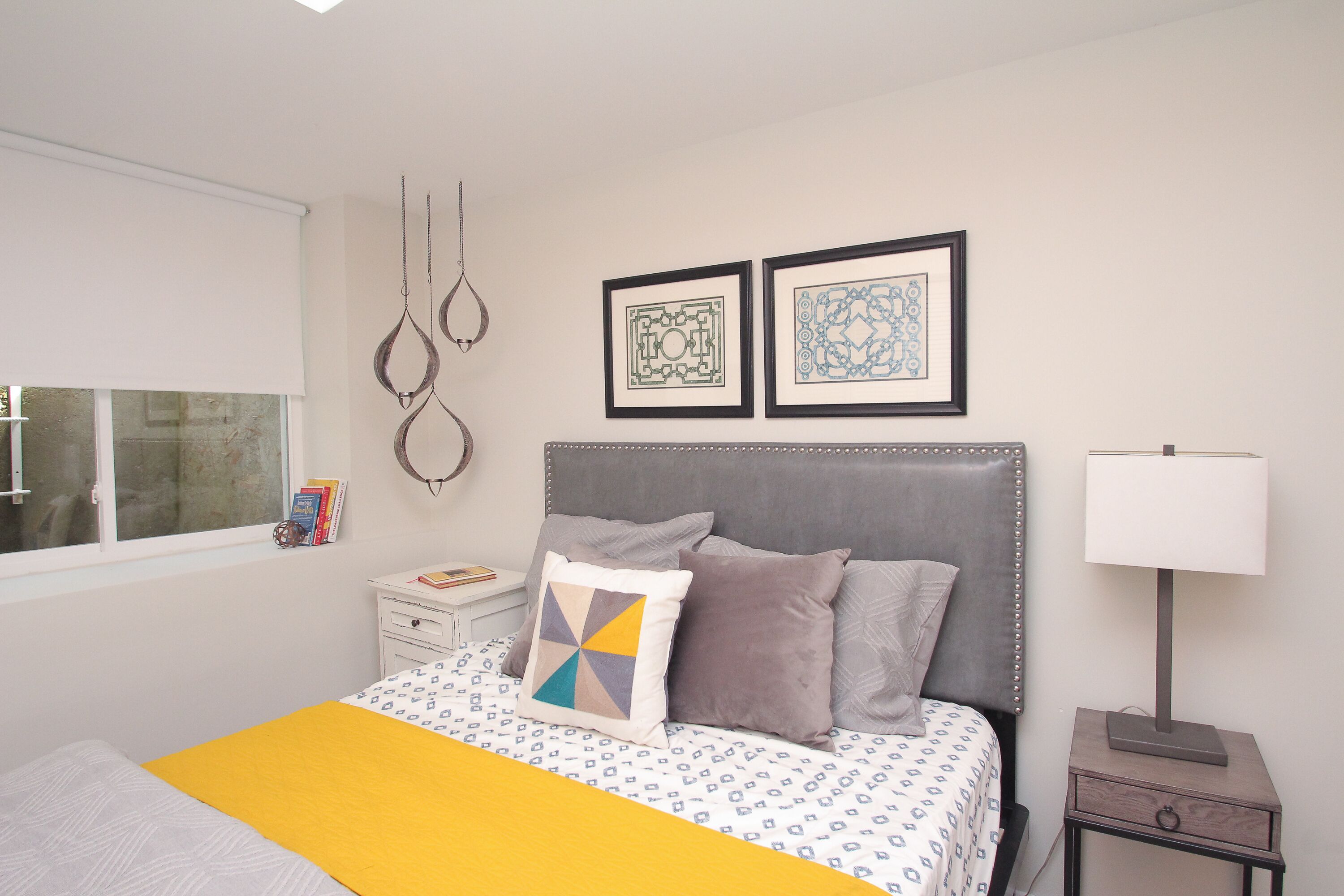 A bedroom with a bed, a bedside table, a nightstand, a window, and two framed pictures on the wall.