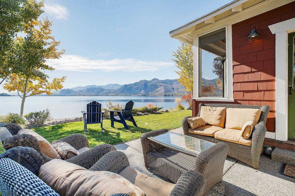 A patio with wicker furniture, a glass table, and a view of a lake and mountains.