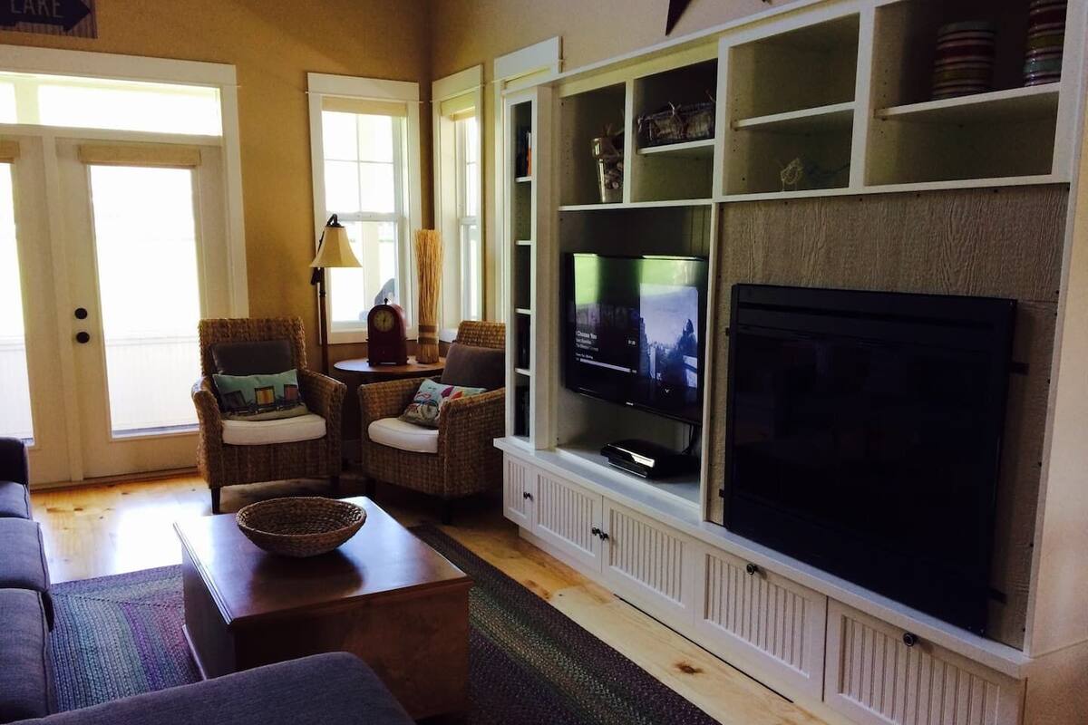 A living room with a large flat-screen TV, wicker chairs, a coffee table, and a fireplace.