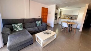 A living room with a grey sectional sofa, a white coffee table, and a dining area with a wooden table and white chairs. - Apartment Los Molinos (Puerto de Naos)
