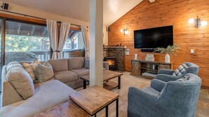 A cozy living room with a sofa, armchairs, and a coffee table. A fireplace is visible in the background, and a flat-screen TV is mounted on the wall.