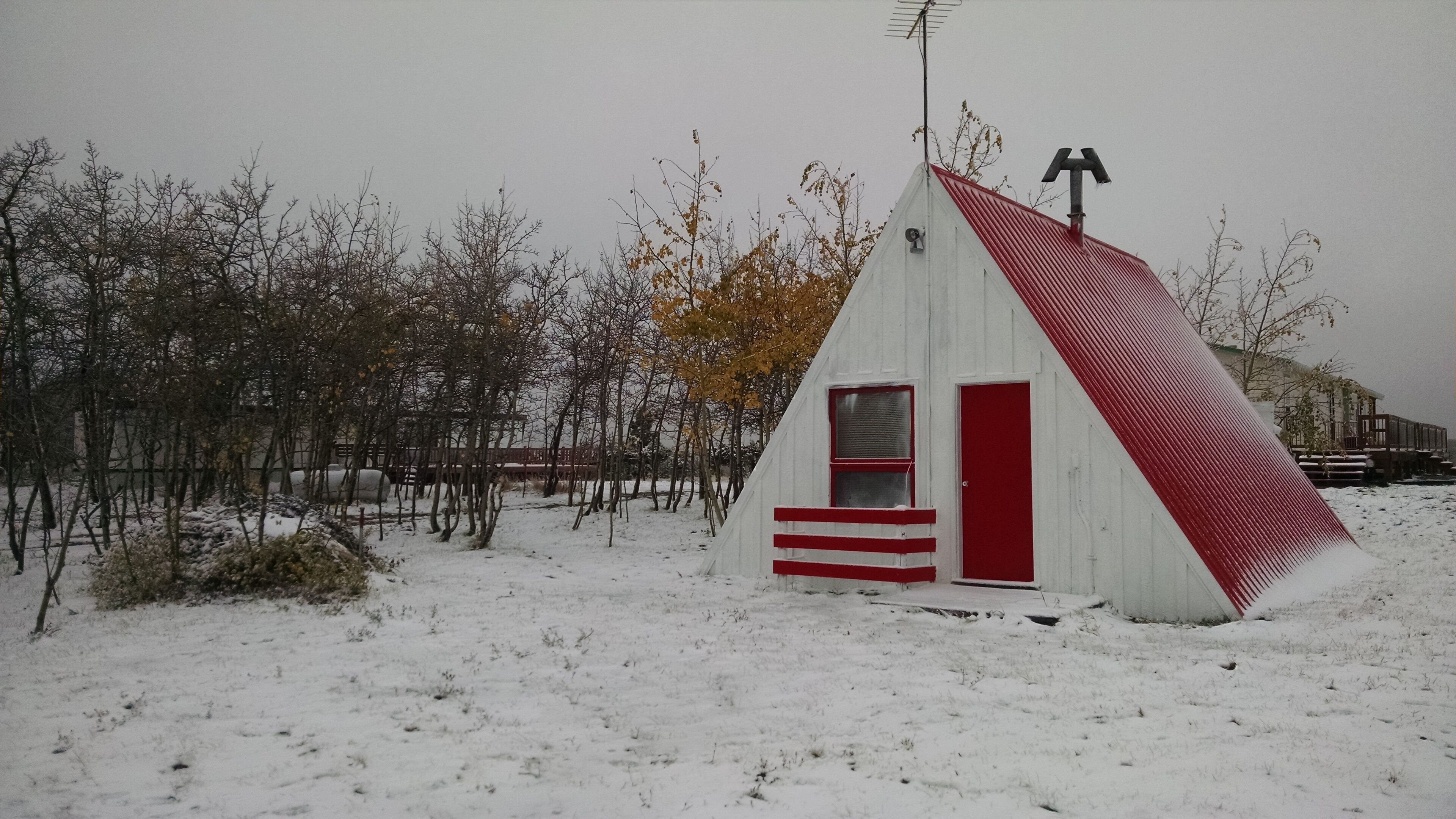 Een klein, wit en rood blokhutje met een schuin dak, gelegen in een besneeuwd landschap met kale bomen.