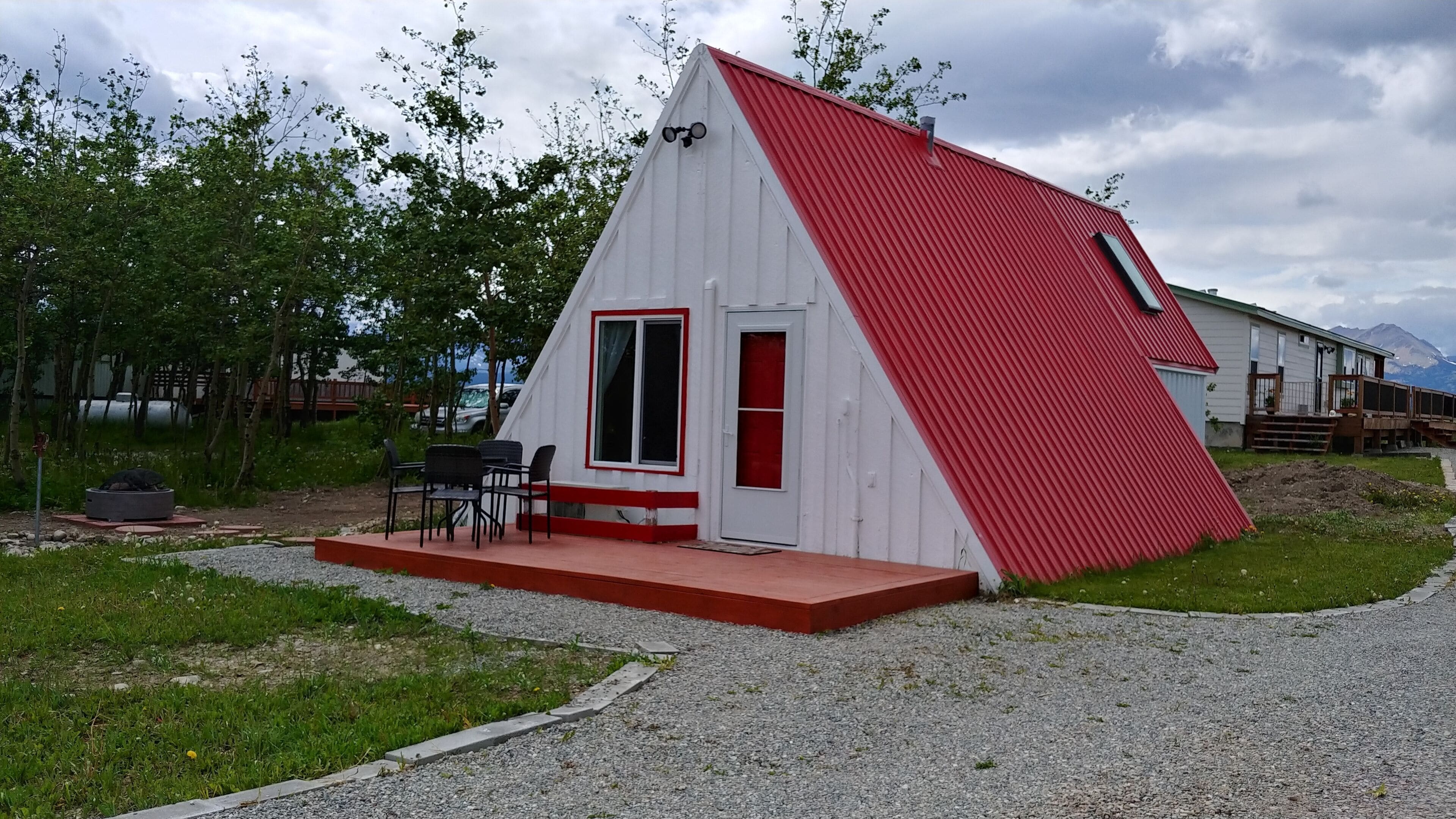Un pequeño edificio triangular blanco y rojo con puerta y ventanas, un patio con sillas y un sendero de grava.