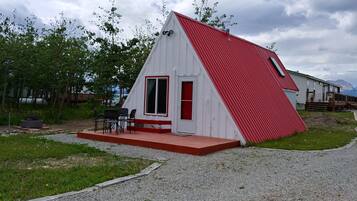 Un pequeño edificio triangular blanco y rojo con puerta y ventanas, un patio con sillas y un sendero de grava.