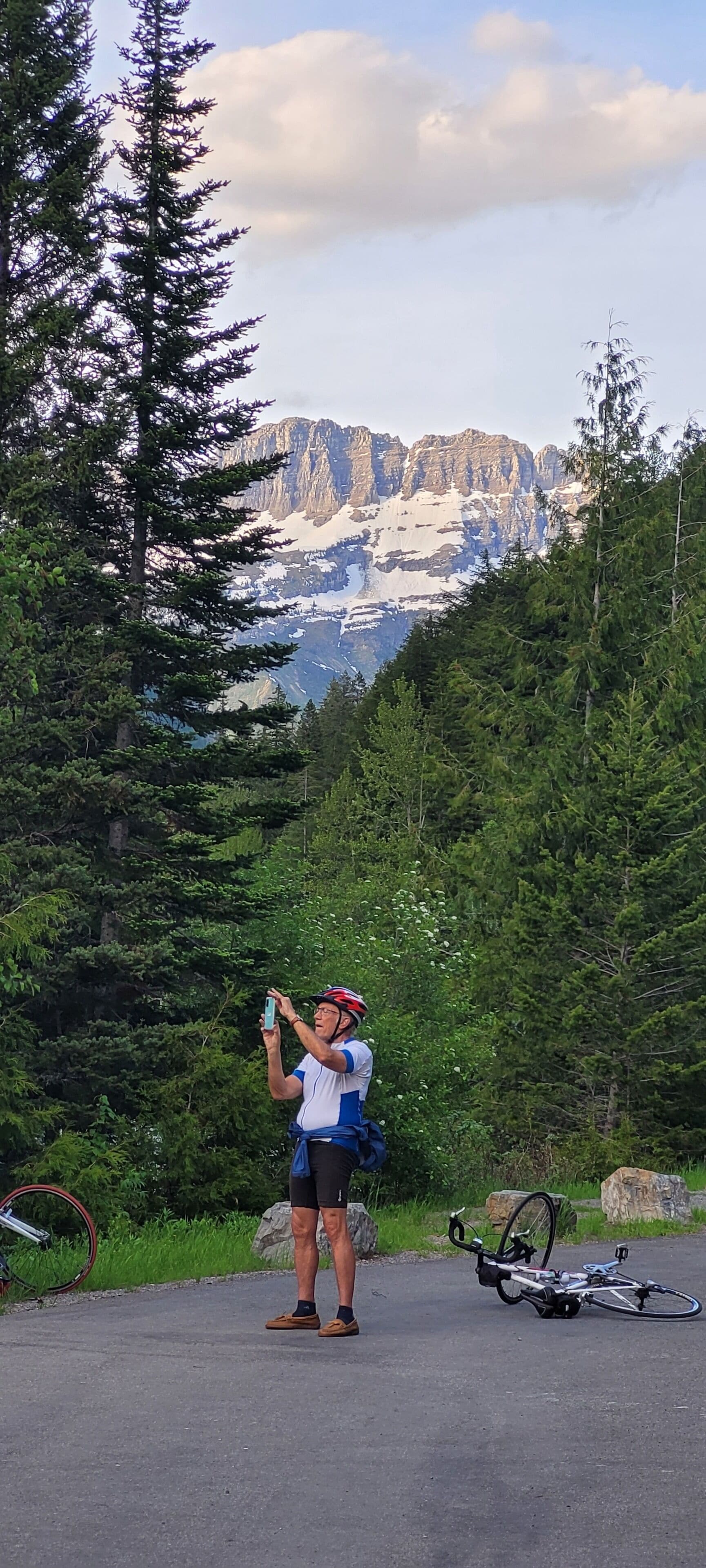 A person taking a photo with a smartphone, standing next to a bicycle on a paved path surrounded by trees and mountains in the background.
