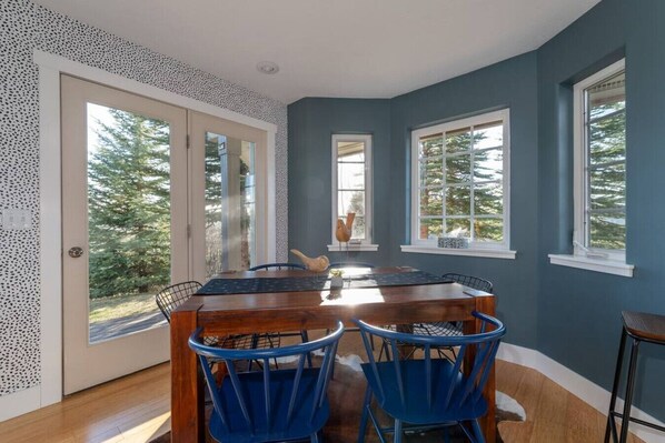A dining area with a wooden table and blue chairs, large windows, and a view of trees.
