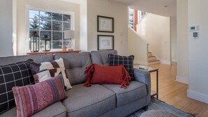 A modern living room with a grey sofa, multiple cushions, a small side table, and a staircase leading to another floor. - Après Chalet – Luxury Town Home off Ski Hill Rd (Driggs)