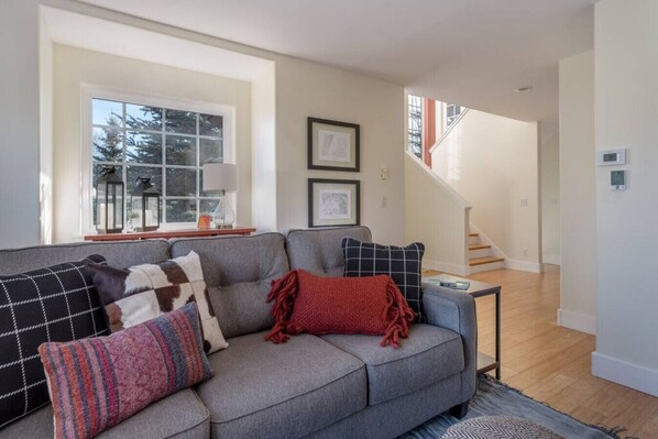 A modern living room with a grey sofa, multiple cushions, a small side table, and a staircase leading to another floor.