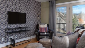 A living room with a patterned wallpaper, a black leather chair, a grey sofa, a flat-screen TV mounted on the wall, and a balcony with a view of buildings and trees. - Après Chalet – Luxury Town Home off Ski Hill Rd (Driggs)