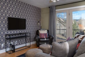 A living room with a patterned wallpaper, a black leather chair, a grey sofa, a flat-screen TV mounted on the wall, and a balcony with a view of buildings and trees.