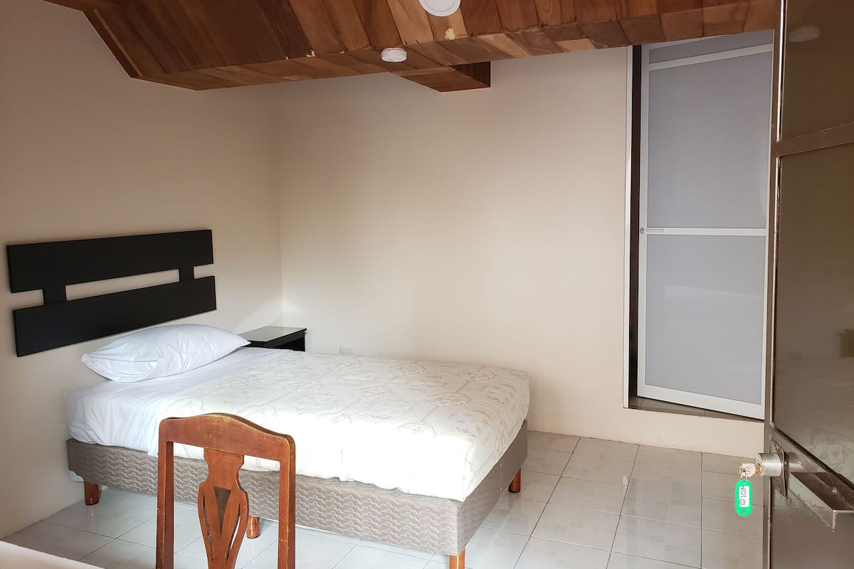 Un dormitorio con cama, una silla de madera, una mesita de noche y una ventana con vistas.