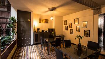 Un comedor moderno con una mesa de vidrio, sillas negras y suelo estampado. Se aprecia una zona de cocina con una cafetera y un reloj en la pared.