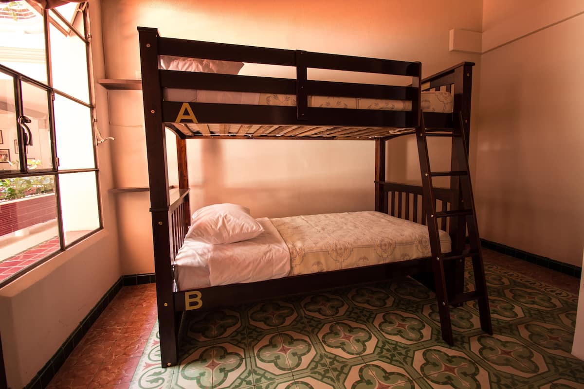 Una cama individual sobre otra con escalera, una ventana con vista a un muro de ladrillo y vegetación, y baldosas de suelo con diseño.